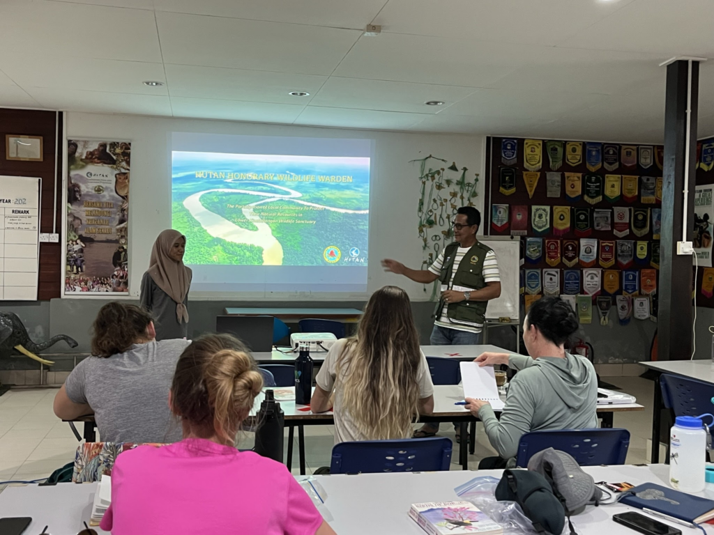 A classroom setting where several adult learners sit at desks facing the front of the room. Two presenters are standing next to a projector screen that is displaying an aerial photograph of a winding river surrounded by a lush, green tropical forest.