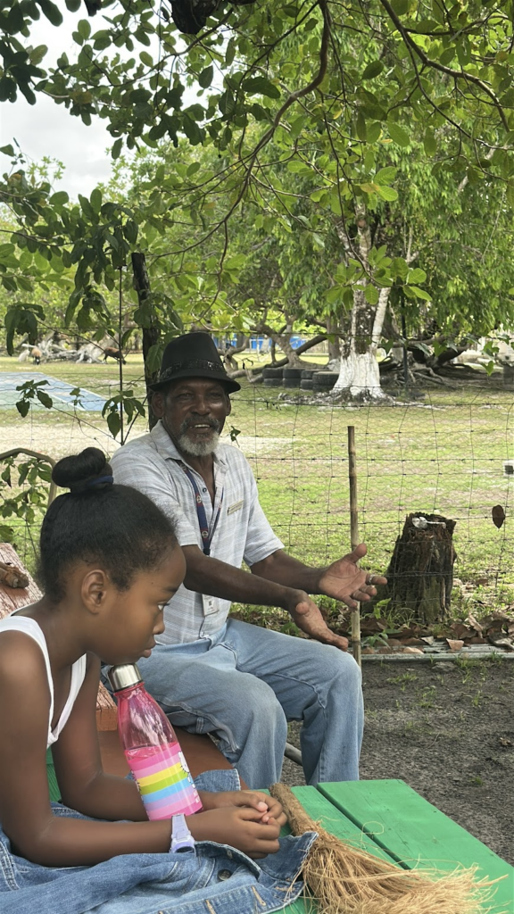 An older Black man with a grey beard, wearing a black fedora hat, a striped short-sleeve button-up shirt, and blue jeans, sits outdoors and gestures with open hands while talking. In the foreground, a young Black girl with her hair in a bun rests her chin on her hand at a green picnic table, looking thoughtfully to the side. A pink water bottle and a straw broom rest on the table beside her. They are seated in a grassy yard shaded by large green trees, with a wire fence in the background.