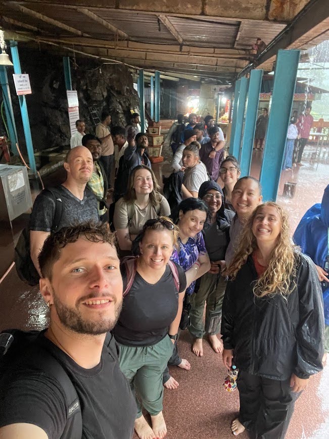 A high-angle selfie captures a group of diverse, smiling students standing together in a long line under a covered walkway during a heavy rainstorm. In the foreground, a man with a short beard and a black t-shirt smiles directly into the camera, leading a group of about nine other travelers who are wearing casual clothing like t-shirts and rain jackets. Several group members in the front are barefoot on the smooth, wet, reddish-brown floor.
Bright blue metal support beams run along the right side of the open-air pavilion, where sheets of rainwater pour down from the roof. In the background, the queue extends deeper into a rocky, sheltered area where local vendors and other people are visible near small stalls under the low wooden ceiling.
