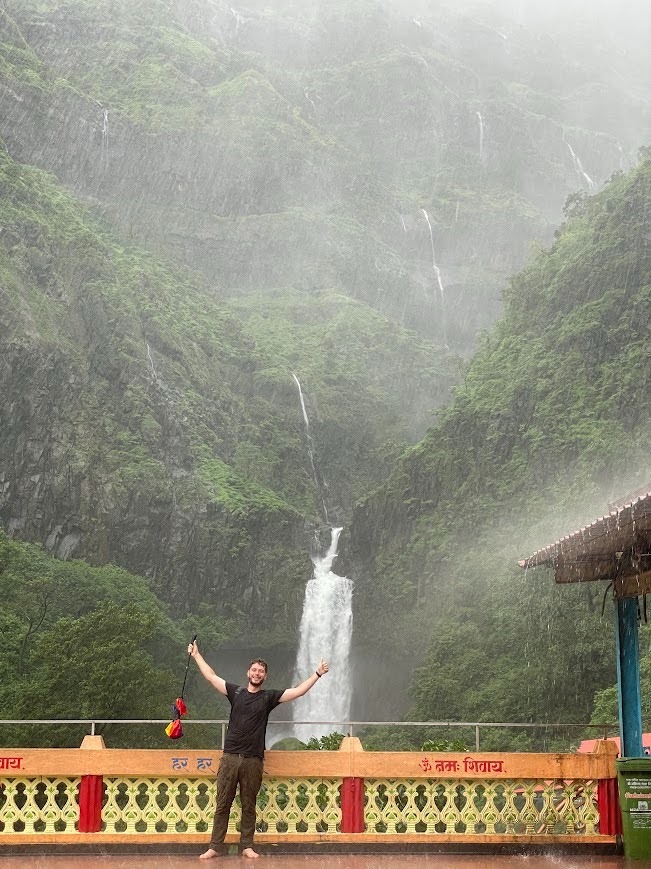 Jack Schaefer stands barefoot on a wet, covered platform with his arms raised wide in celebration during a heavy downpour. He is wearing a dark, rain-soaked t-shirt and pants, holding a closed, colorful rainbow umbrella in one hand.
Behind him, a low decorative railing with red and yellow accents features the Hindi script "हर हर" and "ॐ नमः शिवाय". In the background, a massive, steep mountain covered in lush green vegetation rises into the mist. A large, powerful white waterfall cascades down the center of the mountain, surrounded by several smaller ribbons of waterfalls trickling down the rocky cliffs above it.
