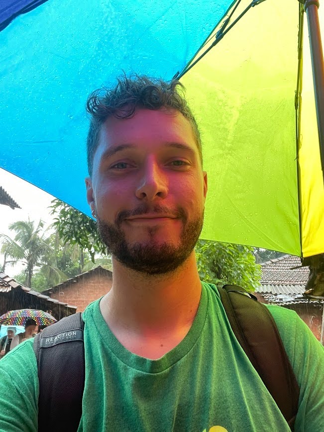 A close-up selfie of Jack Schaefer standing outdoors in the rain. He is wearing a green t-shirt and a black backpack, holding a large rainbow umbrella above him, with its blue and lime-green panels visible directly overhead. The colored panels cast a vibrant hue onto his face. In the background, rain falls over a rural village setting with palm trees, green foliage, small houses with tiled roofs, and another person holding a colorful umbrella in the distance.