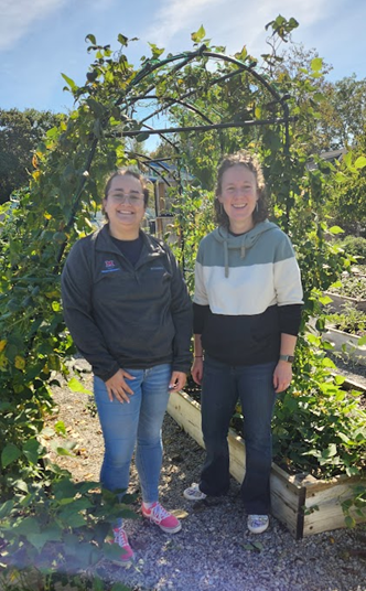 Two smiling professionals stand outdoors under a tall, vine-covered garden trellis on a sunny day. They are surrounded by lush raised planter beds and gravel pathways, and one is wearing a grey Miami University quarter-zip pullover.