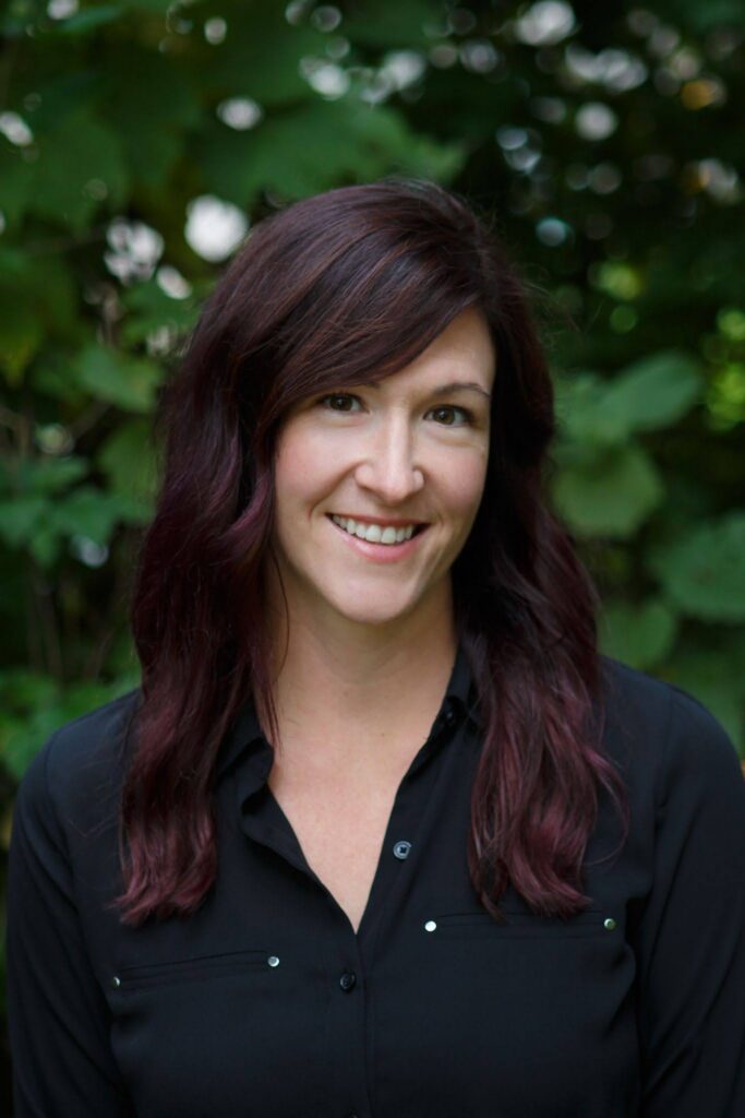 A headshot of Christy Frank smiling, with a blurred background of green foliage.