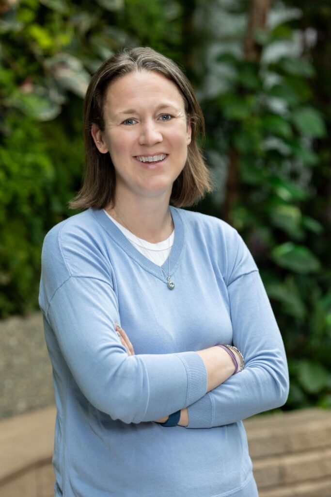 Dr. Claire Lannoye-Hall standing outside at the Detroit Zoo with her arms crossed, wearing a blue sweater and smiling warmly at the camera.