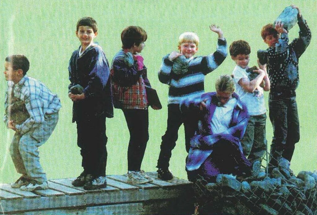 A line of seven elementary and junior high students standing on a wooden platform, smiling and laughing as they lift and pass large rocks down the line.