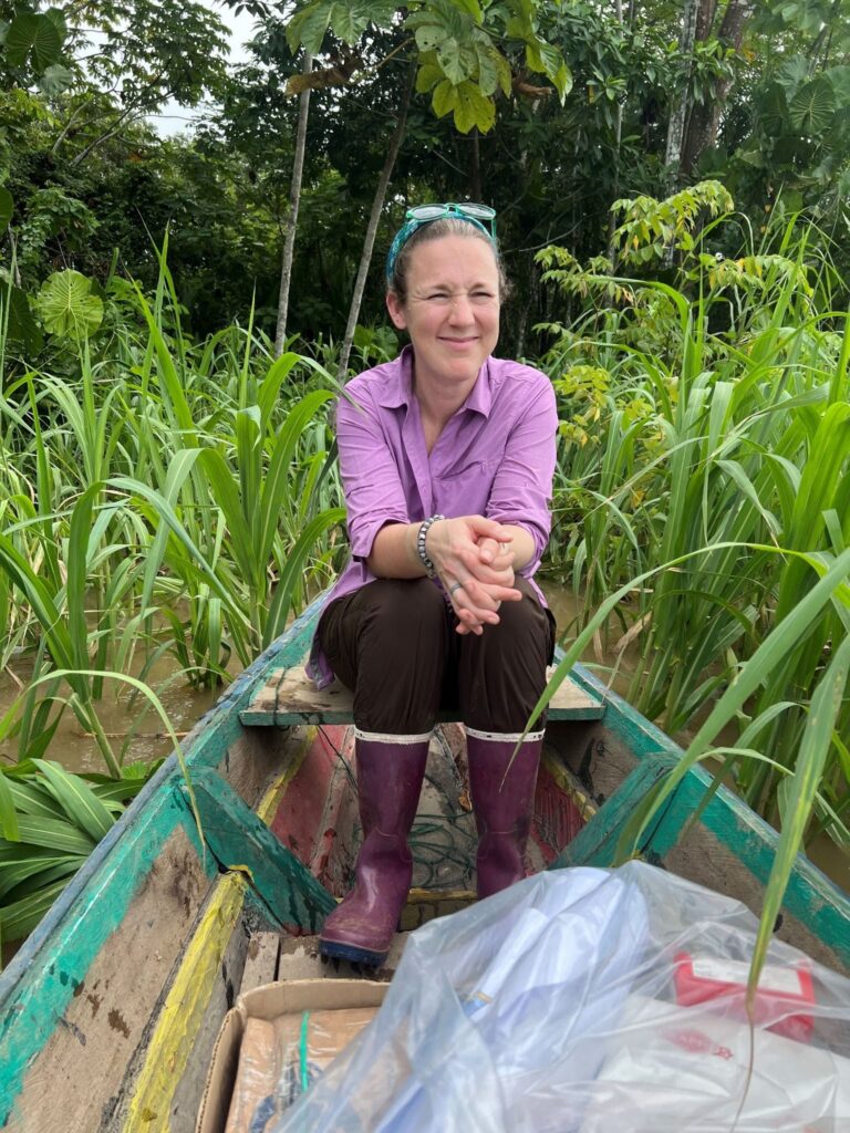 Claire Lannoye-Hall smiling while sitting in a canoe on a river in the Amazon. She has sunglasses resting on her head, tall wading boots on, and thick verdant jungle foliage behind her. 