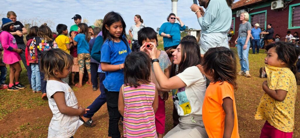 An outdoor gathering with a large group of children and adults on a dirt ground next to a red brick building.

In the center foreground, a woman kneels down to hold a small, orange cylindrical viewing device or toy to a young child's eye, who looks through it curiously. Several other young children stand closely around them, watching intently.

In the background, more children and adults are scattered across the area, chatting, standing in small clusters, and interacting under a slightly overcast sky.