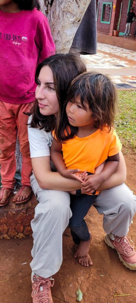 A close-up shot of a Giulia sitting down and holding a young child wearing an orange shirt in her lap. Both are looking off to the left with thoughtful expressions. Another child wearing a pink sweatshirt stands closely next to them on the left, partially captured in the frame. The background shows an outdoor dirt area near the base of a tree.