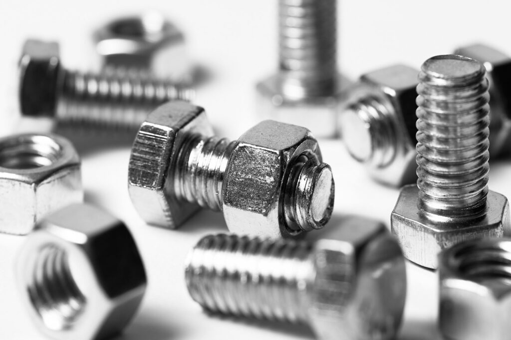 Macro photograph showing a close-up of shiny silver metal bolts with visible spiral threads and hexagonal nuts scattered on a white surface, with one nut screwed onto a bolt in sharp focus.