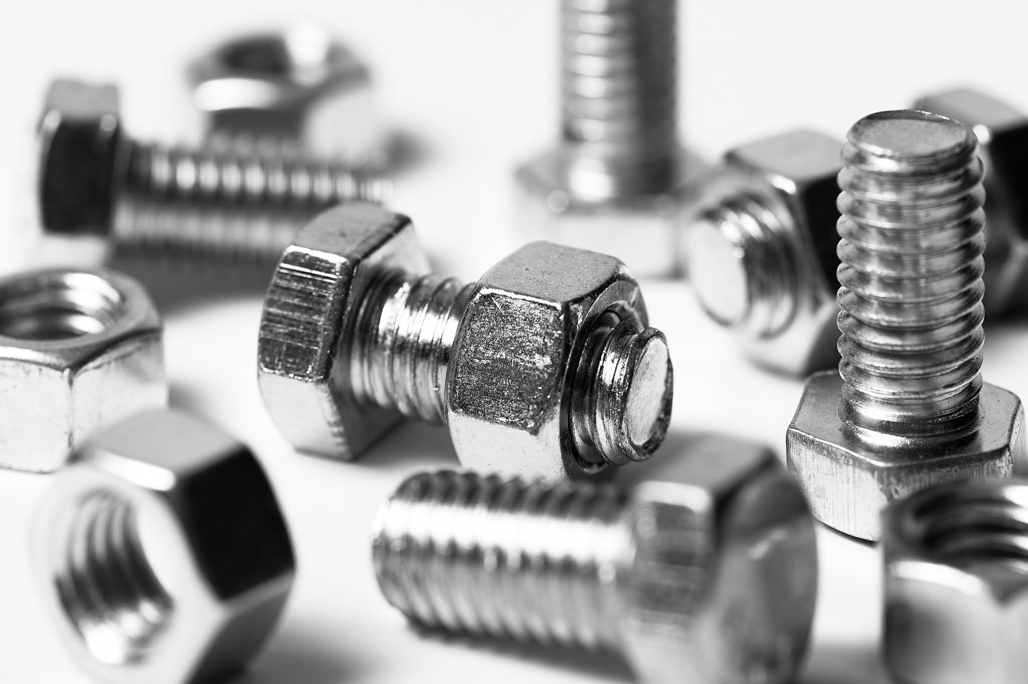 Macro photograph showing a close-up of shiny silver metal bolts with visible spiral threads and hexagonal nuts scattered on a white surface, with one nut screwed onto a bolt in sharp focus.
