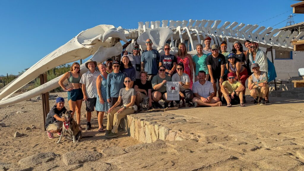 A group of people and a dog pose outdoors in front of a massive whale skeleton, holding a Miami University banner.