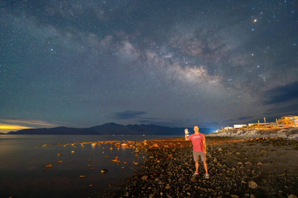 A man waves on a rocky beach at night beneath a bright Milky Way sky with distant mountains.