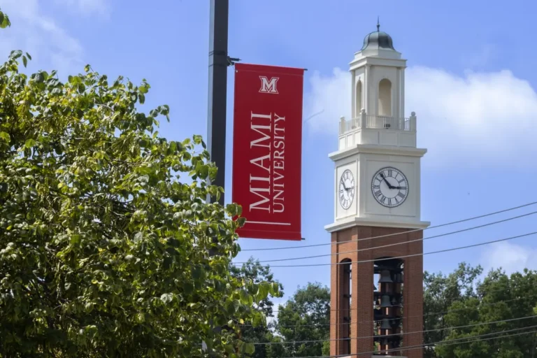 A red banner reading "Miami University" hangs beside a clock tower with a brick base and white upper section, set against a blue sky with green trees.