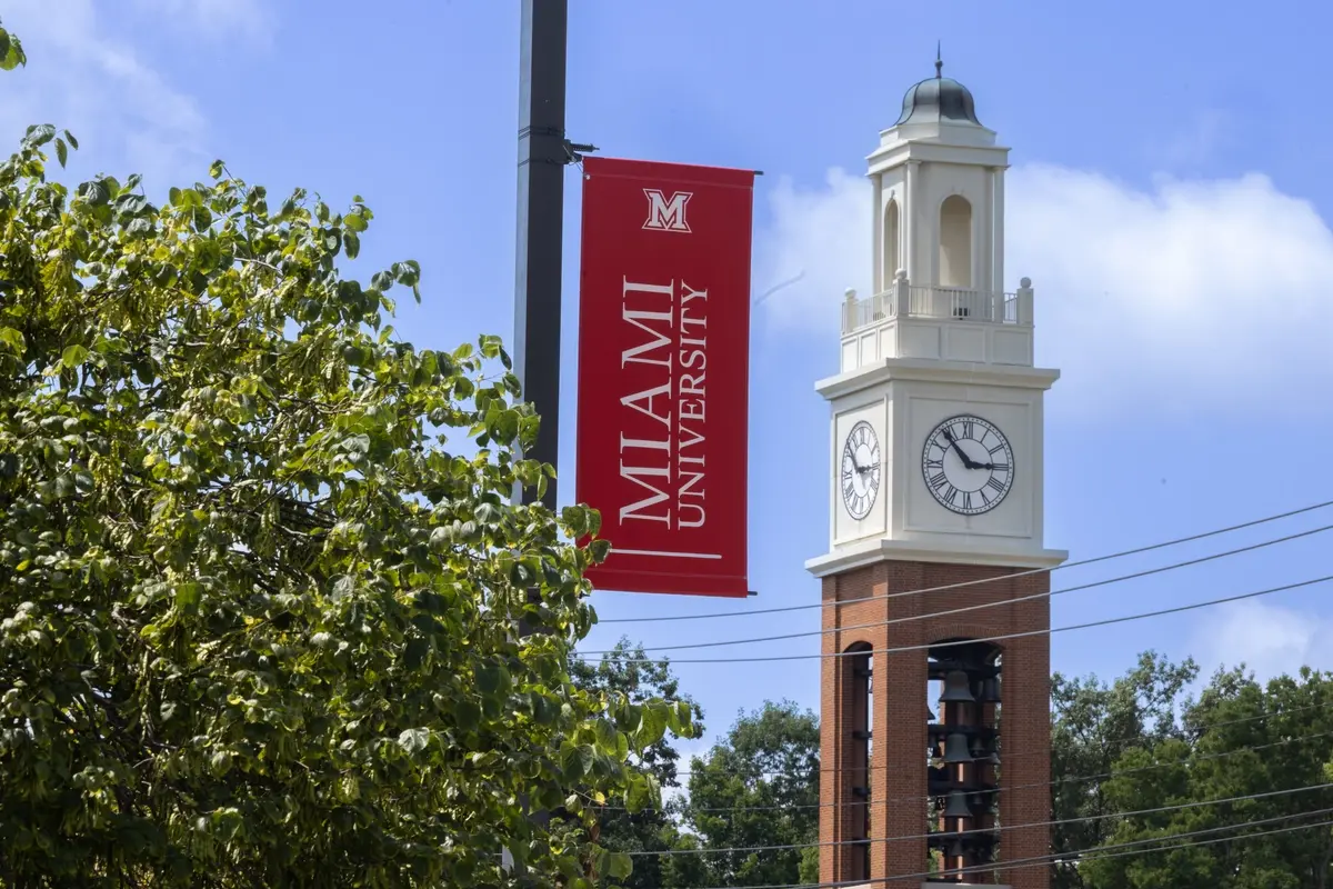 A red banner reading "Miami University" hangs beside a clock tower with a brick base and white upper section, set against a blue sky with green trees.