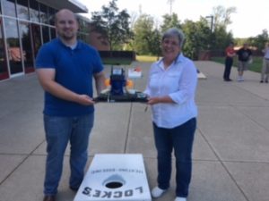 Michael Robinson and Leah Harris holding the IT Services cornhole trophy standing near a cornhole board east of Millett Hall