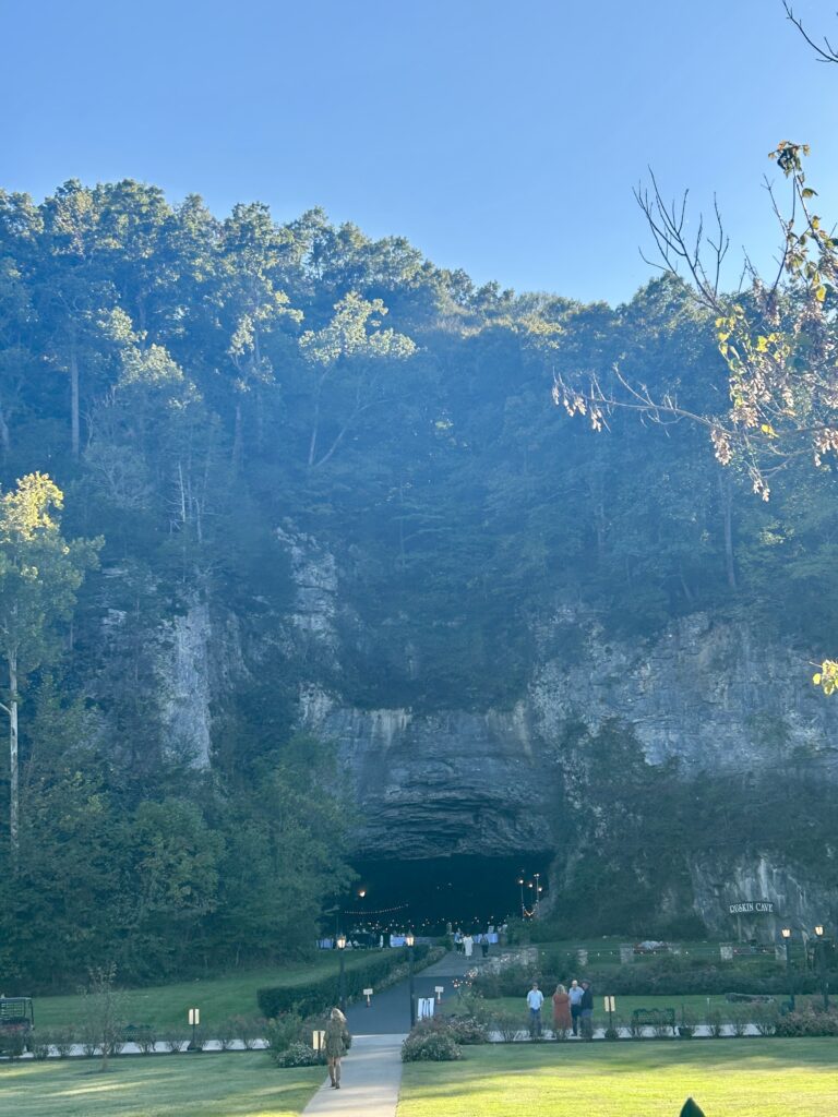 Long-distance shot of the entrance to a cave, where a wedding is taking place. Some people and light fixtures can be seen inside the cave.