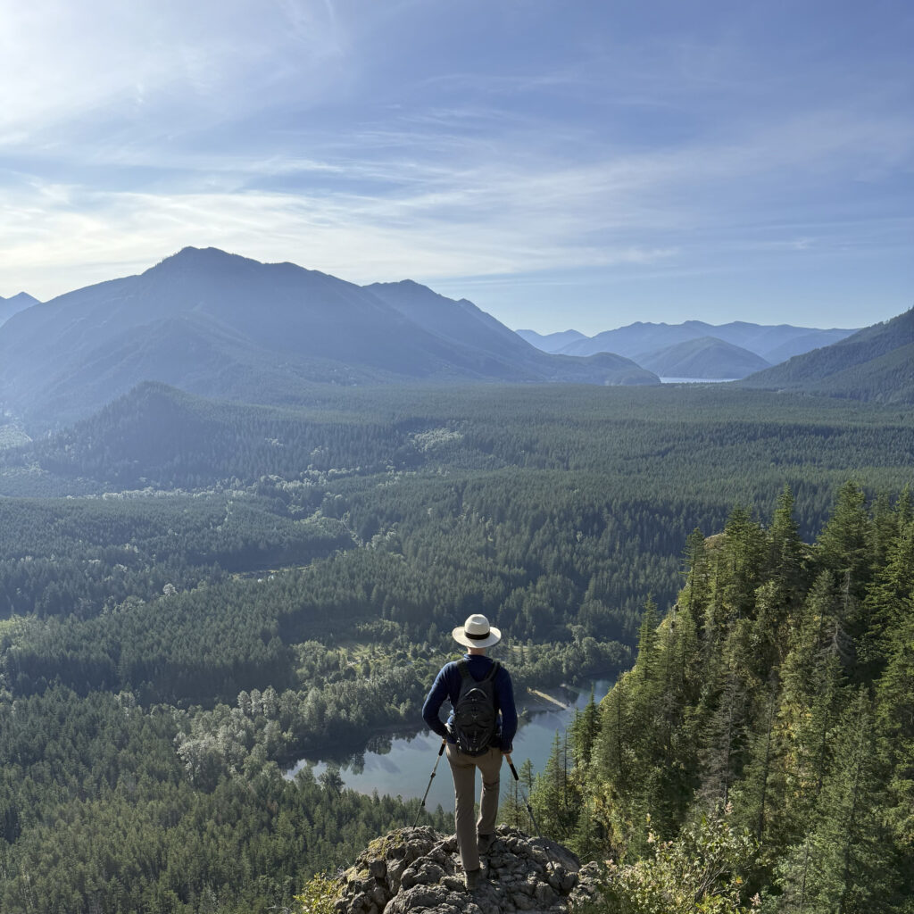a person stands with hiking poles atop a rocky hill, overlooking a landscape of green trees, gentle blue lakes, and towering mountains