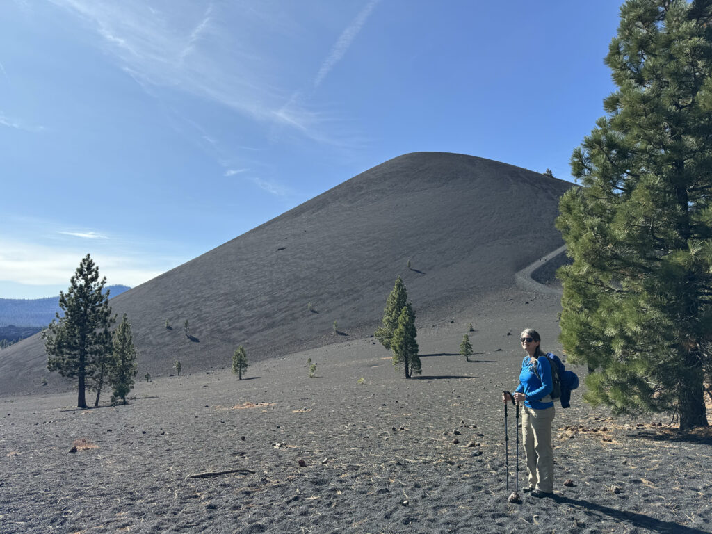 A person stands with hiking poles at the base of a tall, sparsely vegetated, sandy hill. A few evergreen trees dot the landscape around them.