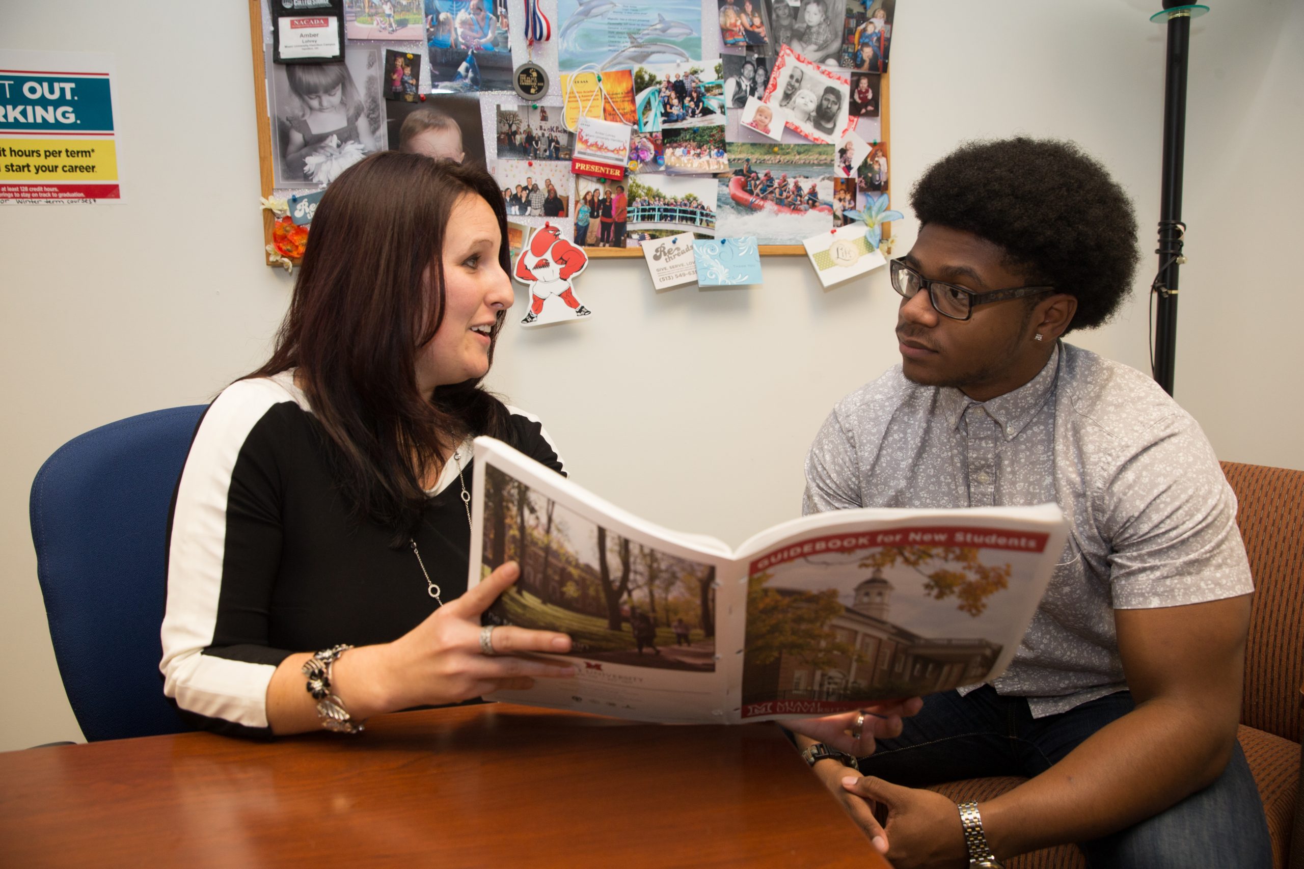 Woman and young man sit next to each other at a table. The woman is holding up an open book and showing the young man something in it.