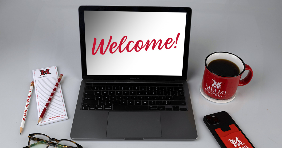 A laptop with "welcome" on the screen, Miami University-branded pencils, a writing list pad, mobile phone wallet, and a cup of coffee.