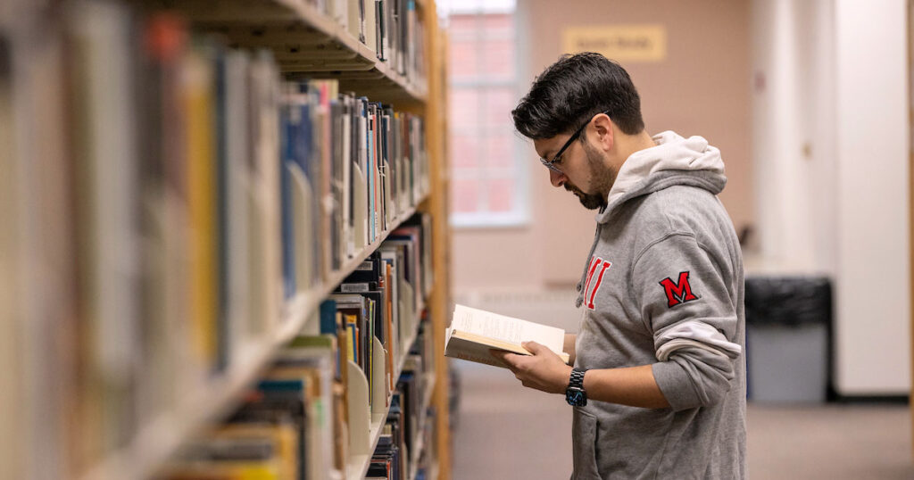 A Miami Online graduate student holding a textbook open at the library