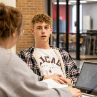 Two students talking having a conversation. One student is typing on a laptop computer.