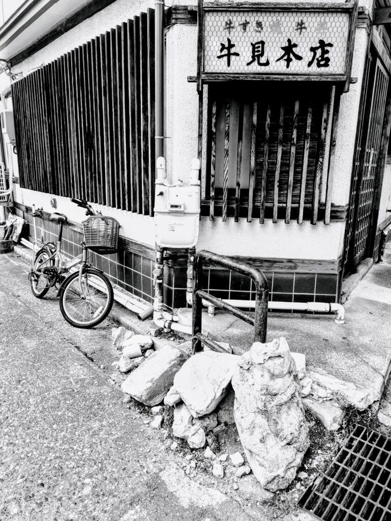 A black and white photo of a run-down street corner in the city of Kyoto. There is a bicycle with a front-basket leaning against a tiled wall that transitions into a wall of wooden bars. There is a honeycomb tiled white sign on the building with Japanese Kanji characters in black lettering.