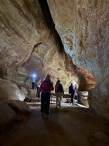 The interior of a cave with twisty rock formations, the background lit by daylight and getting darker towards the foreground. Move Beyond Surviving (MBS) women are eclipsed by the daylight, delving deeper into the cave with a flashlight.