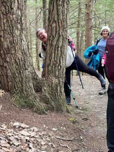 A woman from the Move beyond Surviving (MBS) group posing playfully behind a tree, with more MBS women smiling in the background. Behind them is a wooded area with green trees.