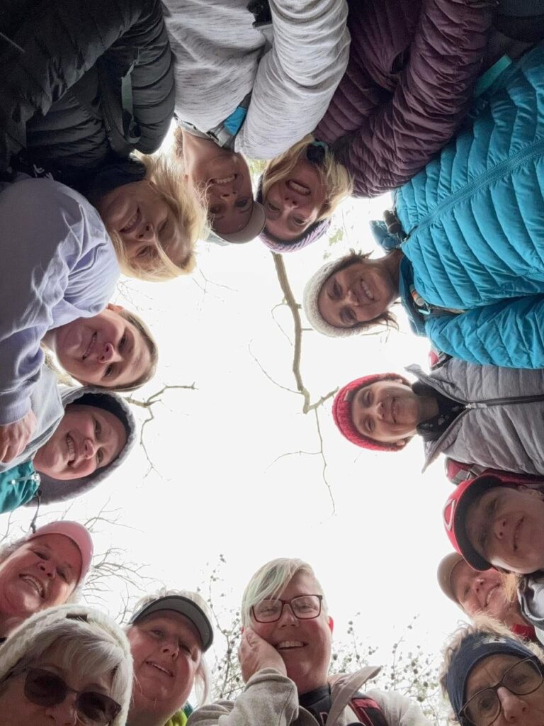 Move Beyond Surviving (MBS) women posed in a circle, smiling down at a camera that captures them from a worms eye view. Daylight and branches are seen overhead in the background.
