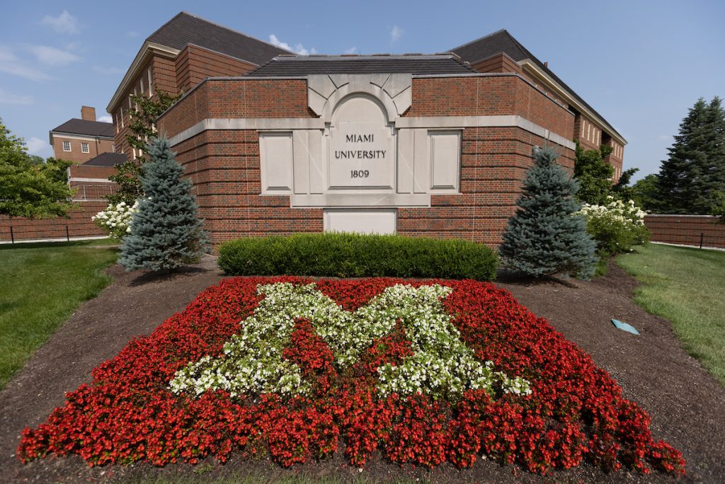 Pearson Hall with the sign that says Miami University 1809. Red and white flowers in the shape of an M in the foreground.