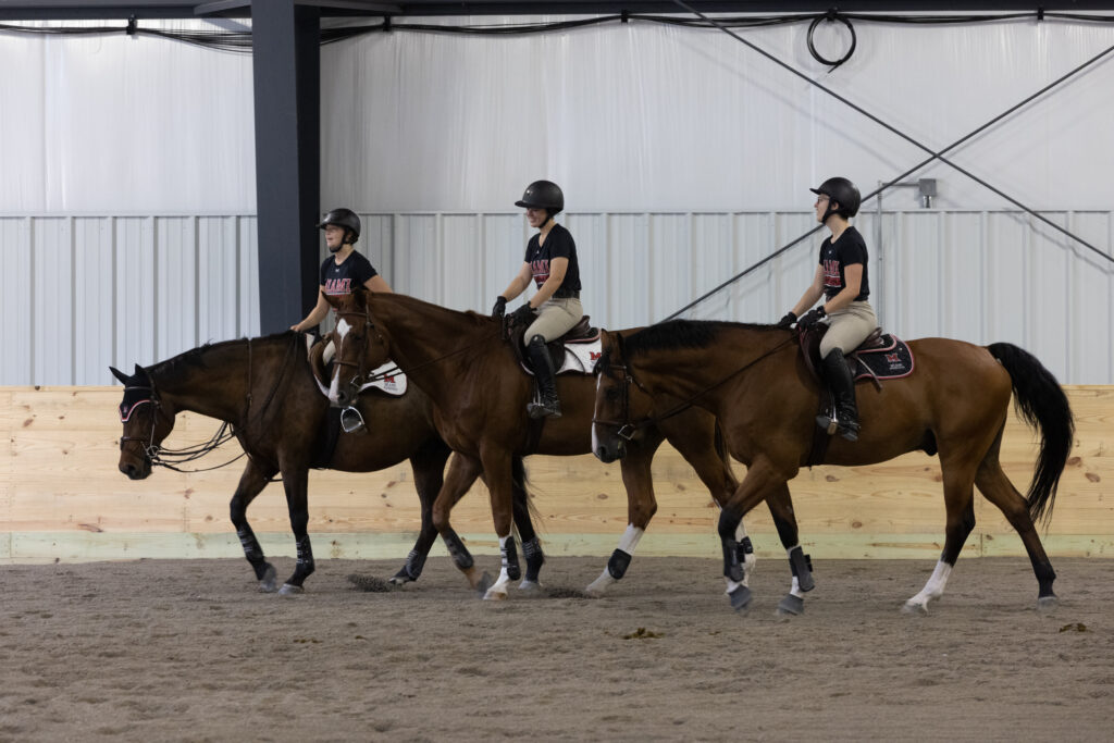 Three women in Miami University gear riding horses in an indoor arena.
