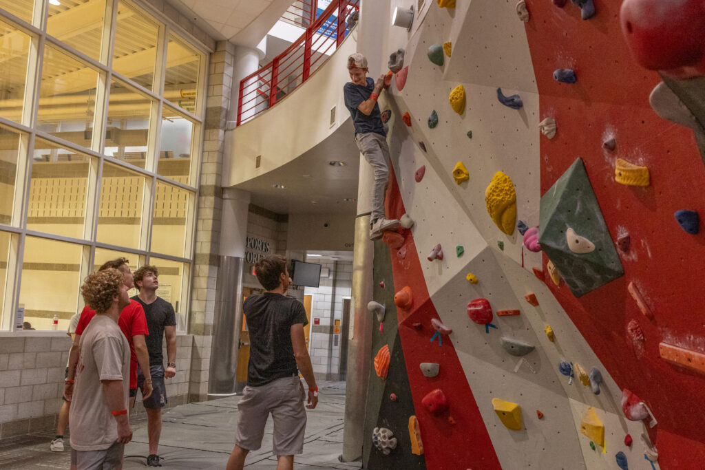 Man climbing indoor rock climbing wall in a gym while 4 others watch.