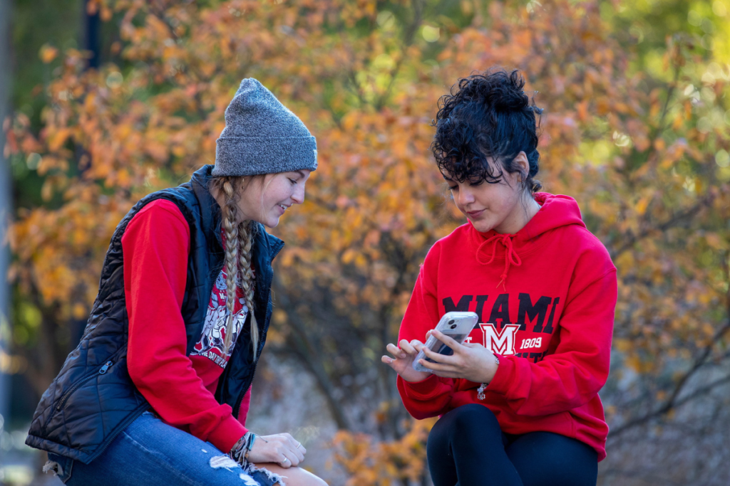 One girl in Miami gear showing another girl something on her phone with trees in the background.